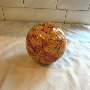 May include: A decorative, spherical object with a glossy finish. The surface is covered in a mosaic of small, irregularly shaped pieces in shades of brown and tan. The object is sitting on a white surface.