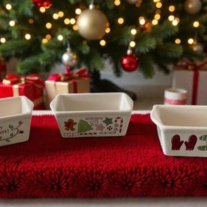 Puede incluir: Tres platos rectangulares de cerámica blanquecina con diseños navideños. Un plato presenta un pingüino y el texto "Happy Holidays!". Los otros dos tienen gráficos festivos como hombres de jengibre, árboles y mitones. Están sobre una superficie roja texturizada.