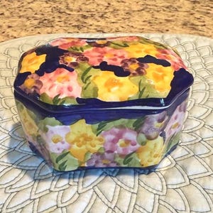 以下が含まれることがあります： 蓋付きの八角形のセラミック製の小物入れ。 黄色、ピンク、紫の色合いの花柄が、濃い青色の背景に描かれています。 白いキルト加工された表面に置かれています。