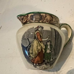 May include: A cream-colored ceramic creamer featuring a green rim and handle. The body displays a detailed illustration of a person in a yellow apron and red skirt standing next to a child in green clothing, with a decorative brown border at the top.