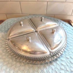 May include: A silver-toned serving dish with four triangular compartments, each with a lid and a small knob. The dish has a decorative beaded edge and is set on a light blue patterned surface. Ideal for serving appetizers or snacks.