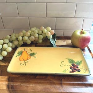 Puede incluir: Una fuente rectangular de cerámica amarilla con diseños de frutas pintados a mano. La fuente presenta un diseño de pera y uvas. Un racimo de uvas y una manzana están al lado de la fuente.