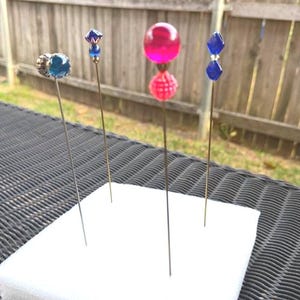 Puede incluir: Una colección de alfileres decorativos para sombreros con varios diseños de cuentas coloridas. Los alfileres presentan cuentas azules, rosas y transparentes en diferentes formas, incluyendo esferas y gemas facetadas. Los alfileres se exhiben sobre una superficie blanca.