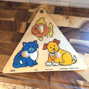 Puede incluir: Rompecabezas triangular de madera con un pez, un gato y un perro. El pez es naranja y amarillo, el gato es azul y blanco, y el perro es amarillo y marrón. Las piezas tienen pomos de madera. La palabra "PETS" está impresa.