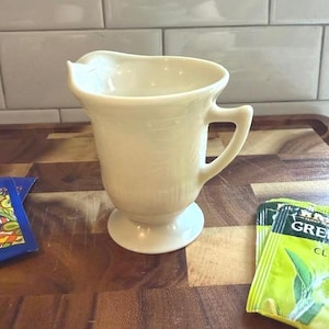 Puede incluir: Cremera de cerámica color crema con asa y base de pedestal. La cremera tiene un pico curvo y una superficie texturizada. La cremera está sobre una superficie de madera. Se ven bolsitas de té en la imagen.
