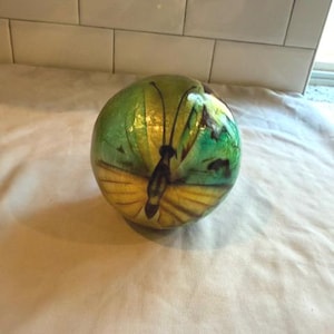 May include: A decorative, round object with a butterfly design. The object is primarily green and yellow, with a black butterfly silhouette. The butterfly's wings are yellow with green accents, and the body is black. The object is sitting on a white surface.