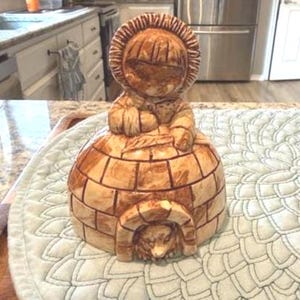 Op de afbeelding: Een keramieken beeldje van een persoon in een parka met capuchon die op een iglo zit. Het beeldje is geschilderd in bruin- en beigetinten, met de iglo met een gedetailleerd baksteenpatroon en een gewelfde deuropening met een berengezicht.