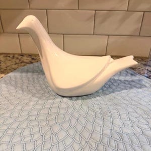 May include: A white ceramic dove figurine with a smooth, glossy finish. The bird is perched on a light blue, patterned placemat. The dove has a long neck and a slightly open beak, suggesting a peaceful pose. The background features a tiled wall.