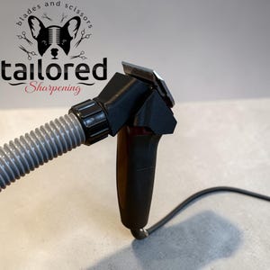 May include: A black electric hair clipper with a metallic blade, attached to a grey flexible vacuum hose. The device features a black power cord and a logo at the top reading blades and scissors tailored Sharpening with a dog illustration.