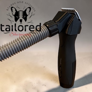 May include: A black electric clipper with a metallic blade head attached to a flexible grey ribbed hose. The device features a power button and indicator lights on the handle. A logo with a dog silhouette and the text tailored Sharpening is visible.