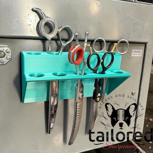 May include: A teal scissor holder with various barber shears and scissors. The holder is mounted on a gray metal surface. The image includes the text "blades and scissors" and "tailored sharpening" with a dog graphic.
