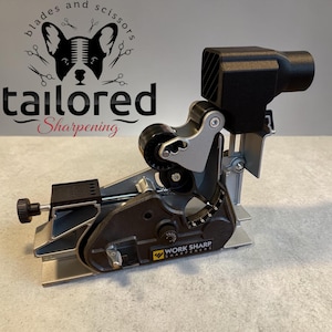 May include: A Work Sharp knife sharpener with a black and silver design. The sharpener has a black handle and a silver base with a dial. The text "Work Sharp" is visible on the side. The image also includes the text "blades and scissors" and "Tailored Sharpening".