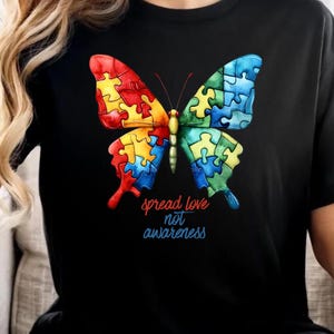 May include: Black t-shirt featuring a colorful butterfly design. The butterfly wings are composed of puzzle pieces in red, yellow, blue, and green. The text "spread love not awareness" is below the butterfly.