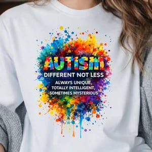 Könnte beinhalten: Weißes T-Shirt mit farbenfrohem Grafikdesign. Das Design zeigt das Wort "AUTISMUS" in Regenbogenfarben, umgeben von einem lebendigen Farbspritzer. Darunter stehen die Worte "ANDERS NICHT WENIGER" und weitere positive Aussagen.