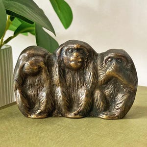 May include: A bronze-colored sculpture of the three wise monkeys: see no evil, hear no evil, speak no evil. The monkeys are sitting side-by-side, with their hands covering their eyes, ears, and mouth. The sculpture is on a green surface.
