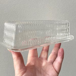 K&ouml;nnte beinhalten: Eine transparente, rechteckige Butterdose aus Kunststoff mit einem strukturierten, geometrischen Muster auf dem Deckel. Die Dose wird in einer Hand vor einem neutralen Hintergrund gehalten. Der Deckel hat eine Reihe vertikaler Rippen und rautenf&ouml;rmiger Facetten.