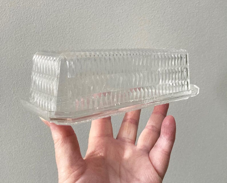 K&ouml;nnte beinhalten: Eine transparente, rechteckige Butterdose aus Kunststoff mit einem strukturierten, geometrischen Muster auf dem Deckel. Die Dose wird in einer Hand vor einem neutralen Hintergrund gehalten. Der Deckel hat eine Reihe vertikaler Rippen und rautenf&ouml;rmiger Facetten.
