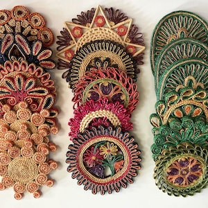 May include: A collection of decorative coasters in various colors, including orange, red, green, and purple. The coasters are made of woven material and feature intricate floral and geometric designs. The coasters are arranged in stacks and rows, showcasing their unique patterns.