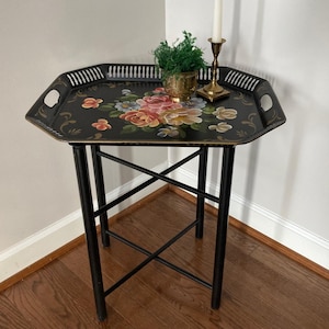 May include: Black, octagonal serving tray table with floral design. The tray has a black background with a colorful floral arrangement in the center. The table has black legs and a decorative edge.