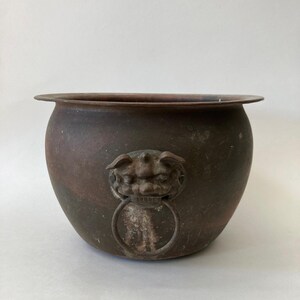 May include: A round, dark bronze-toned metal bowl featuring a wide, flat rim and a weathered, matte finish. The exterior is adorned with a sculpted lion head ornament that holds a circular metal ring, providing a decorative handle detail.