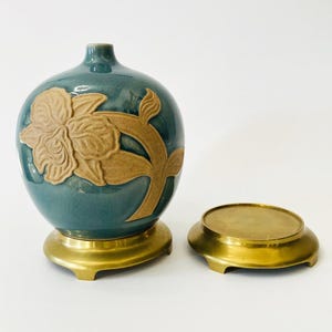 May include: A teal ceramic vase with a floral design in tan, set on a gold-colored base. Beside it is a matching gold-colored saucer. The vase has a rounded shape and a narrow neck. The floral design is embossed.
