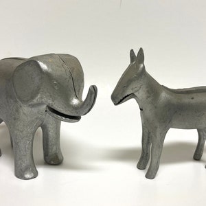 May include: Two gray metal coin banks: an elephant and a donkey. The elephant has a trunk and a slot on top. The donkey has a long snout and a slot on its back. Both are standing on a white surface.