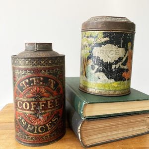 May include: Two antique metal canisters. One is labeled "TEA COFFEE SPICES" in red and black. The other, with a lid, has a black background with a painted scene and the word "RICE". Both are on top of green books.