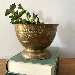 May include: A polished brass bowl with a pedestal base, featuring an embossed floral pattern around the center and decorative scalloped bands along the top and bottom rims. The bowl contains a small green plant with several oval-shaped leaves.