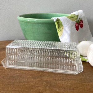 K&ouml;nnte beinhalten: Eine klare, rechteckige Butterdose aus Glas mit einem Rautenmuster. Eine gr&uuml;ne Keramiksch&uuml;ssel, ein wei&szlig;es und gr&uuml;n gemustertes Geschirrtuch mit roten Erdbeeren und zwei wei&szlig;e Eier befinden sich im Hintergrund. Die Objekte stehen auf einer Holzoberfl&auml;che.