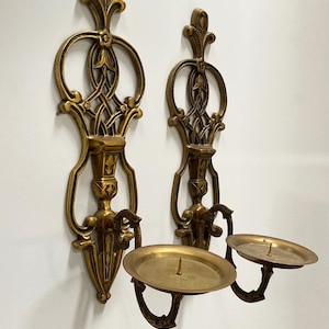 May include: Two ornate, antique-style wall sconces in a warm gold tone. Each sconce features a decorative backplate with intricate scrollwork and a circular candle holder. The sconces are designed to hold candles and add a touch of elegance.