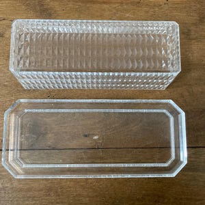 K&ouml;nnte beinhalten: Eine transparente, rechteckige Glasbox mit einem strukturierten Deckel mit Rautenmuster. Die Basis ist ein transparenter, rechteckiger Tablett mit achteckigem Rand. Die Artikel werden auf einer Holzoberfl&auml;che pr&auml;sentiert.