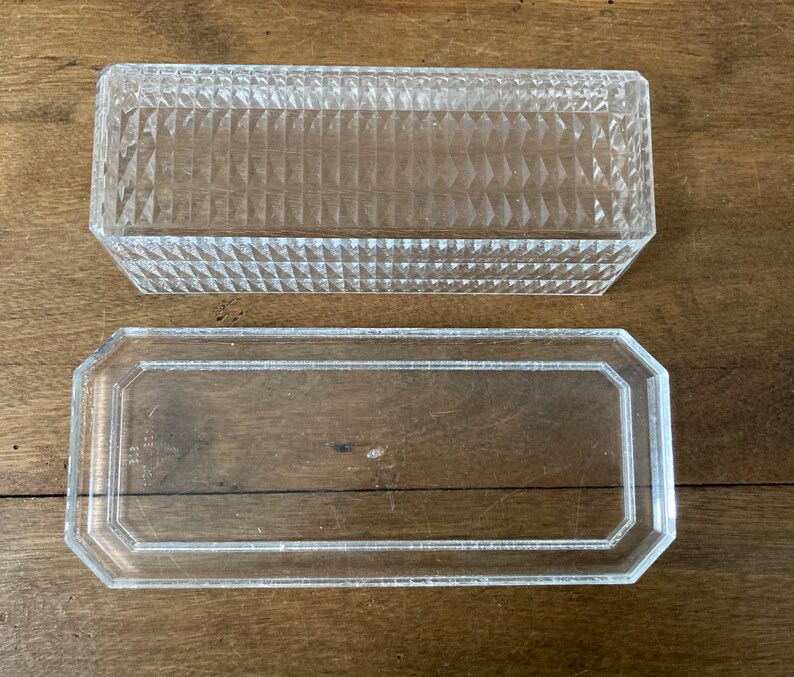 K&ouml;nnte beinhalten: Eine transparente, rechteckige Glasbox mit einem strukturierten Deckel mit Rautenmuster. Die Basis ist ein transparenter, rechteckiger Tablett mit achteckigem Rand. Die Artikel werden auf einer Holzoberfl&auml;che pr&auml;sentiert.