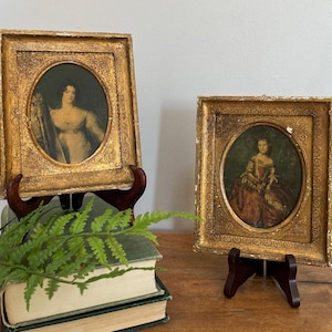 May include: Two rectangular gold frames with oval openings, each containing a portrait of a person. The frames feature ornate, textured detailing and are displayed on dark brown stands. One frame rests on a stack of two books.