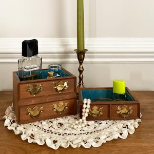 May include: Two small wooden dresser-style boxes with brass hardware and teal interiors. One box has two drawers, the other has one. A perfume bottle, a small jar, and a green-capped bottle sit inside the boxes. A green candle and a pearl necklace are also present.