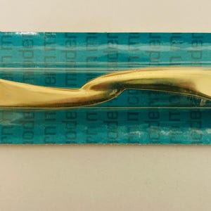 May include: A gold-colored metal cabinet pull with a smooth, curved, and elongated S-shape design. The hardware is encased in clear plastic packaging attached to a teal-colored cardboard backing that features the repeated text modern modern.