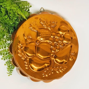 May include: A vintage copper-colored metal mold with a scalloped edge. The mold features embossed images of grapes, wheat, and other fruit. A small loop is attached for hanging. The mold is round and has a shiny finish.