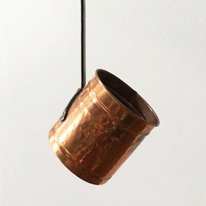 May include: A copper-colored, hammered metal pot with a dark handle. The pot has a cylindrical shape with a slightly angled rim. The handle is attached to the side of the pot with a small metal bracket. The background is a plain white.