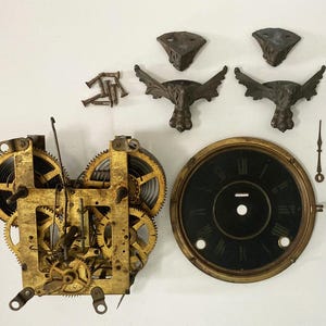 May include: Disassembled antique clock parts, including a brass movement with gears and springs, a black clock face with Roman numerals, decorative feet, and hands. The components are arranged on a white surface.