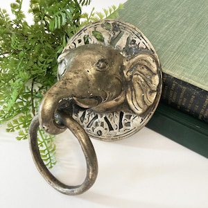 May include: Antique brass elephant door knocker with a circular backplate and a ring for knocking. The elephant head is detailed with wrinkles and a trunk. The knocker is set against a white surface with green foliage and a green book in the background.