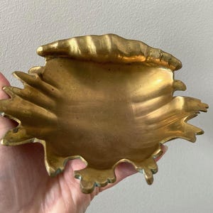 May include: A gold-colored metal dish with a decorative, scalloped edge. The dish is shaped like a seashell and has a smooth, shiny surface.