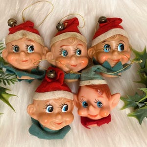 May include: A collection of vintage Christmas elf head ornaments. Each ornament features a smiling elf face with blue eyes, a red hat, and a green or red collar. The ornaments have gold string hangers and small bells.