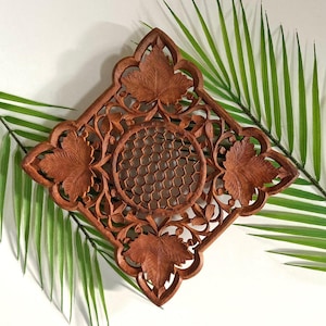 May include: A square, wooden wall decor piece with intricate carvings. The design features four large leaves at the corners, a central honeycomb pattern, and floral accents. The wood has a warm, reddish-brown hue.