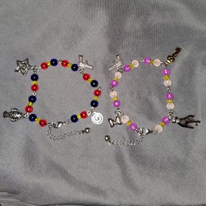 May include: Two beaded bracelets with silver-tone metal charms. The left bracelet features red and blue beads with a star, fish, and pistol charm. The right bracelet has pink and purple beads with a pistol, bow, key, and hand charm.