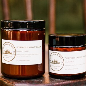 May include: Two glass jars of whipped tallow balm with black lids and white labels. The larger jar is labeled "lavender + vanilla" and the smaller jar is labeled "simply tallow". The jars are on a wooden surface.