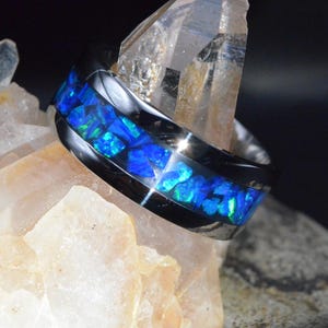 Op de afbeelding: Een zwarte ring met een band van helder blauw en groen opaalachtig materiaal. De ring wordt getoond tegen een achtergrond van lichtgekleurde kristallen, wat een opvallend contrast creëert. Het ontwerp van de ring is modern en opvallend.