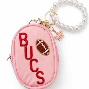 May include: A light pink oval-shaped zippered pouch featuring the red embroidered text BUCS and a brown football patch with gold glitter trim. The pouch includes a gold-toned metal keychain with a white faux pearl wristlet and a GAME DAY charm.