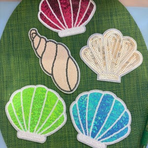 May include: Five decorative patches shaped like seashells. One is dark pink, one is beige, one is light gold, one is bright lime green, and one is turquoise blue. Each patch is covered in small, shimmering sequins with a white border outline.