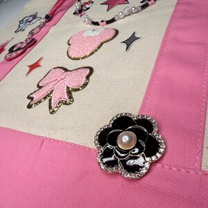 May include: A cream-colored canvas tote bag with pink fabric trim, decorated with a black and white flower brooch featuring a central pearl, a pink glittery bow patch, a pink mouse-ear patch, and a beaded strap with star and pearl accents.