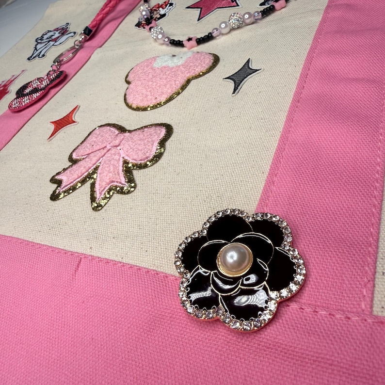 May include: A cream-colored canvas tote bag with pink fabric trim, decorated with a black and white flower brooch featuring a central pearl, a pink glittery bow patch, a pink mouse-ear patch, and a beaded strap with star and pearl accents.