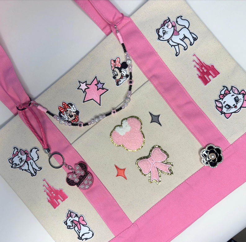 May include: A cream-colored canvas tote bag with pink straps and trim, decorated with embroidered patches of cartoon cats, a castle, stars, and bows. A beaded lanyard with a pink glittery mouse ear keychain is attached to the bag strap.